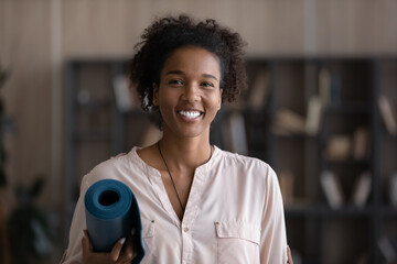 Portrait of smiling young African American woman hold yoga mat ready to practice meditate at home. Happy millennial mixed race female coach or client do sports train stretching or pilates.