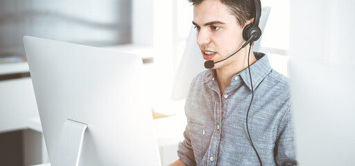 Casual dressed young man using headset and computer while talking with customers online in sunny office. Call center, business concept