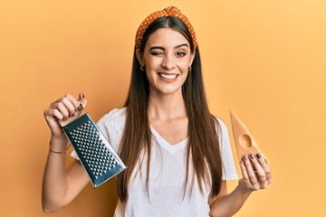 Beautiful brunette young woman holding grater and cheese winking looking at the camera with sexy...