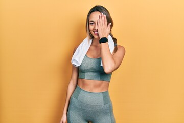 Beautiful hispanic woman wearing sportswear and towel covering one eye with hand, confident smile on face and surprise emotion.