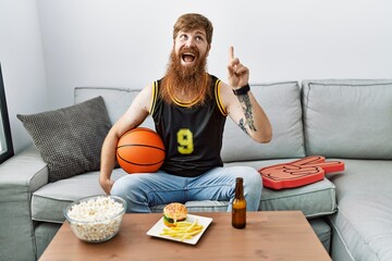 Caucasian man with long beard holding basketball ball cheering tv game smiling amazed and surprised and pointing up with fingers and raised arms.