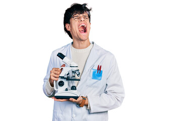 Handsome hipster young man wearing lab coat holding microscope angry and mad screaming frustrated and furious, shouting with anger looking up.