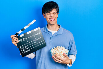 Handsome hipster young man eating popcorn holding cinema clapboard sticking tongue out happy with...