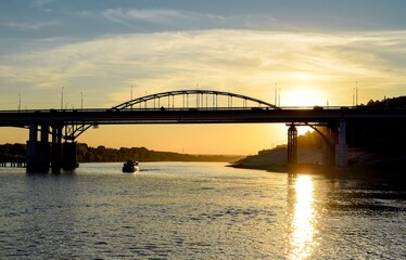 
The silhouette of the bridge over the river and the ship passing under it against the sunset background

