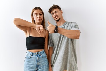 Young beautiful couple standing together over isolated background doing thumbs up and down, disagreement and agreement expression. crazy conflict