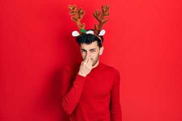 Young hispanic man wearing cute christmas reindeer horns smelling something stinky and disgusting,...