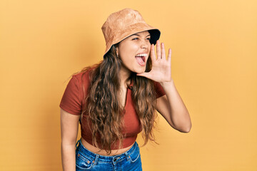 Young hispanic girl wearing casual clothes and hat shouting and screaming loud to side with hand on mouth. communication concept.