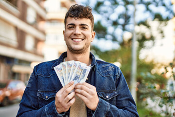 Young hispanic man smiling happy holding peruvian sol banknotes at the city - obrazy, fototapety, plakaty