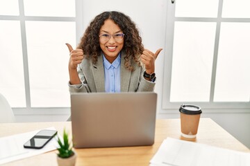 Beautiful hispanic business woman sitting on desk at office working with laptop success sign doing positive gesture with hand, thumbs up smiling and happy. cheerful expression and winner gesture.