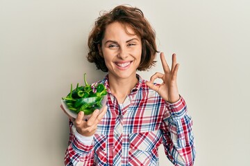 Young brunette woman holding bowl with green peppers doing ok sign with fingers, smiling friendly gesturing excellent symbol