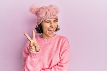 Young brunette woman wearing cute wool cap smiling with happy face winking at the camera doing victory sign. number two.