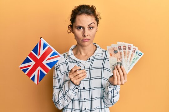 Young brunette woman holding uk flag and pounds banknotes skeptic and nervous, frowning upset because of problem. negative person.