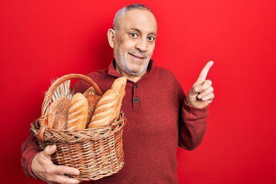 Handsome mature man holding wicker basket with bread smiling happy pointing with hand and finger to the side