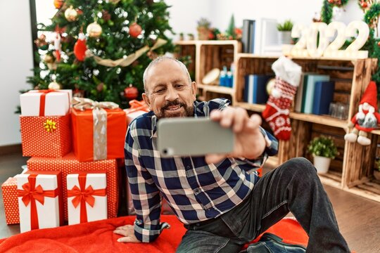 Senior man with beard wearing santa claus hat celebrating christmas at home, sitting on the floor by christmas tree and presents taking a selfie picture with smartphone