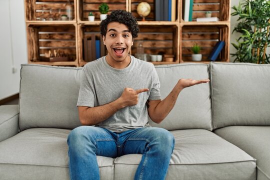 Young hispanic man wearing casual clothes sitting on the sofa at home amazed and smiling to the camera while presenting with hand and pointing with finger.