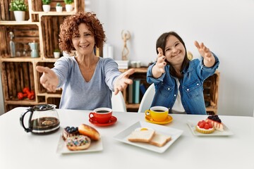 Family of mother and down syndrome daughter sitting at home eating breakfast looking at the camera smiling with open arms for hug. cheerful expression embracing happiness.