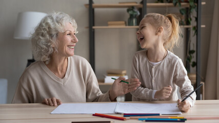 Overjoyed loving old grandmother and small granddaughter have fun drawing painting together in album. Smiling middle-aged Caucasian grandma and little grandchild enjoy hobby art activity at home.