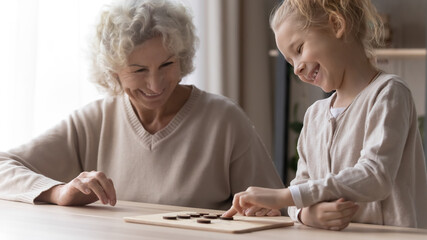 Smiling loving old grandmother and little granddaughter have fun play wooden board game together. Happy mature grandma and small grandchild feel playful with chess or draughts. Hobby concept.