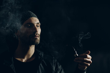Man smokes smoking pipe against dark background. Back side lit profile portrait with smoke