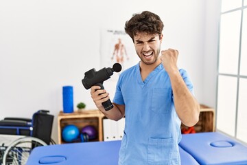 Hispanic physiotherapy man working at pain recovery clinic with muscle gun annoyed and frustrated shouting with anger, yelling crazy with anger and hand raised