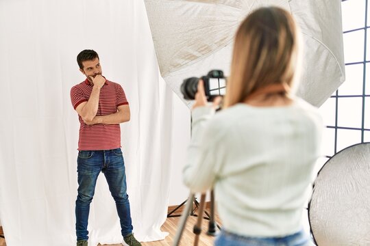 Woman photographer talking pictures of man posing as model at photography studio serious face thinking about question with hand on chin, thoughtful about confusing idea