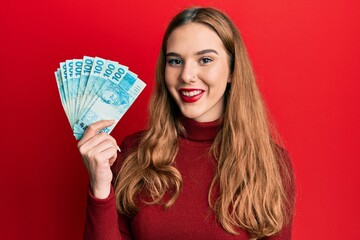 Young blonde woman holding 100 brazilian real banknotes looking positive and happy standing and...