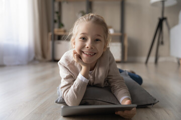 Portrait of happy little teen Caucasian girl child lie relax on floor at home have fun using tablet. Smiling small kid rest talk speak on video call, engaged in webcam virtual event on pad online.