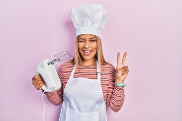Beautiful hispanic woman holding pastry blender electric mixer smiling with happy face winking at...