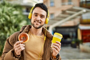 Fototapeta premium Young hispanic man having breakfast using headphones at the city.
