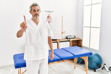 Middle age hispanic therapist man working at pain recovery clinic pointing with finger up and angry expression, showing no gesture