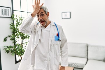 Middle age hispanic man wearing doctor uniform and stethoscope at waiting room surprised with hand...