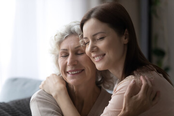 Close up of happy young grownup woman and older 50s mom hug cuddle enjoy close tender family moment together. Smiling adult daughter embrace mature senior mother show love and care. Unity concept.