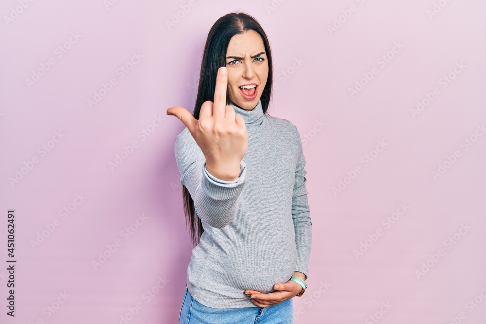 Sticker Beautiful woman with blue eyes expecting a baby, touching pregnant belly showing middle finger, impolite and rude fuck off expression