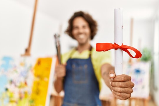 Young hispanic artist man smiling happy holding graduate diploma at art studio.