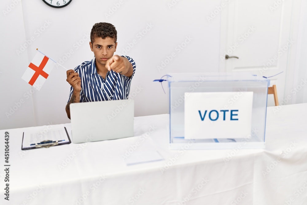 Sticker Young handsome hispanic man at political campaign election holding england flag pointing with finger to the camera and to you, confident gesture looking serious