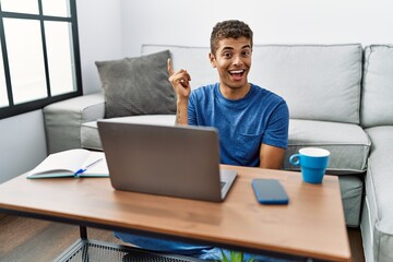Young handsome hispanic man using laptop sitting on the floor with a big smile on face, pointing with hand and finger to the side looking at the camera.