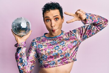 Handsome man wearing make up holding shiny disco ball making fish face with mouth and squinting...