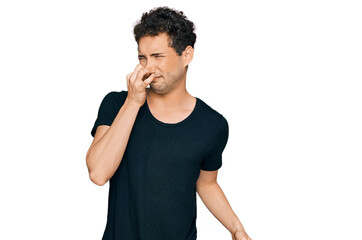 Young handsome man wearing casual black t shirt smelling something stinky and disgusting, intolerable smell, holding breath with fingers on nose. bad smell