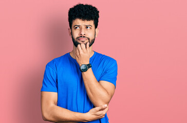 Young arab man with beard wearing casual blue t shirt with hand on chin thinking about question, pensive expression. smiling and thoughtful face. doubt concept.