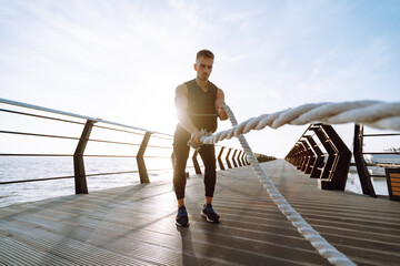 Young man wearing sports clothes is doing exercisesat the beach pier in the morning. Battle ropes....