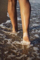 Closeup detail of female legs walking along the seashore. Women's legs and feet walk along the sand and sea waves, along the beach. Relaxation travel concept. Vertical photo.