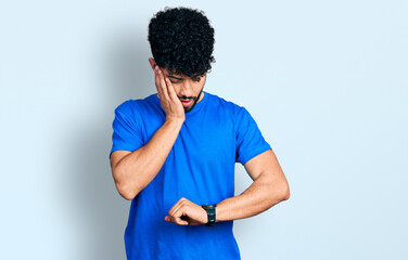 Young arab man with beard wearing casual blue t shirt looking at the watch time worried, afraid of...