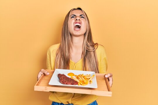 Beautiful hispanic woman holding tray with meat loaf and fried egg angry and mad screaming frustrated and furious, shouting with anger looking up.