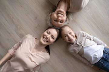 Top view portrait of smiling three generations of Caucasian women lying on wooden floor have fun relax together. Happy little girl child with young mother and old grandmother play show family unity.