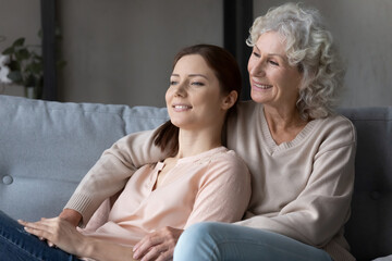 Obraz premium Smiling millennial woman and senior mother relax on sofa at home look in distance dreaming imagining. Happy old mom and adult grownup daughter rest together enjoy family time. Vision concept.