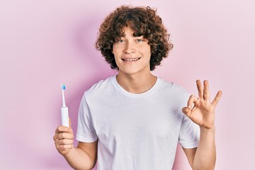 Handsome young man holding electric toothbrush doing ok sign with fingers, smiling friendly gesturing excellent symbol