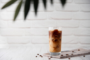 Iced coffee in a tall glass with cream on a white wood background. Cold tasty summer refreshment beverage concept. Selective focus, copyspace.