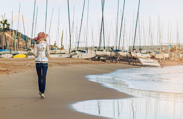 rear view of a Caucasian woman wearing a hat strolling along the shore at sunset.