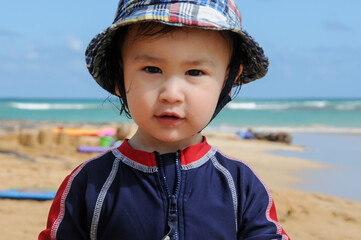Closeup of face of adorable Asian-American toddler boy at the beach wearing plaid hat and swim...