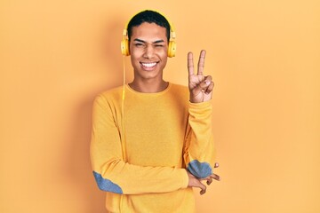 Young african american guy listening to music using headphones smiling with happy face winking at...